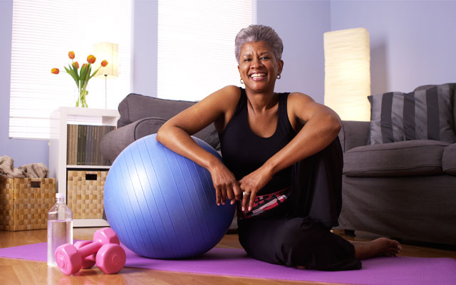 image of woman in living room sitting on yoga mat and leaning on yoga ball with weights and a water bottle close by